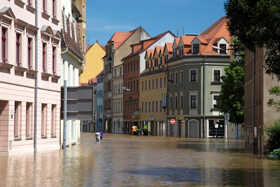 An image of a flood-affected residential area showing partially submerged brick houses with flat roofs, surrounded by muddy water. The water level reaches the lower parts of the buildings, with some s
